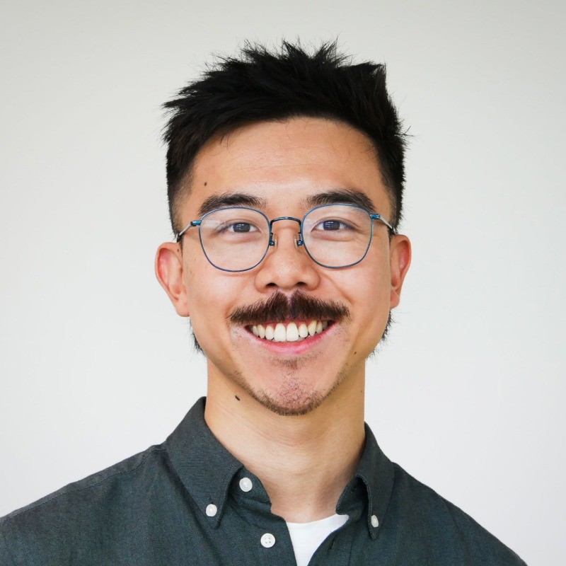 A handsome young man of Chinese descent with a moustache smiles in a dark green collared shirt and glasses.